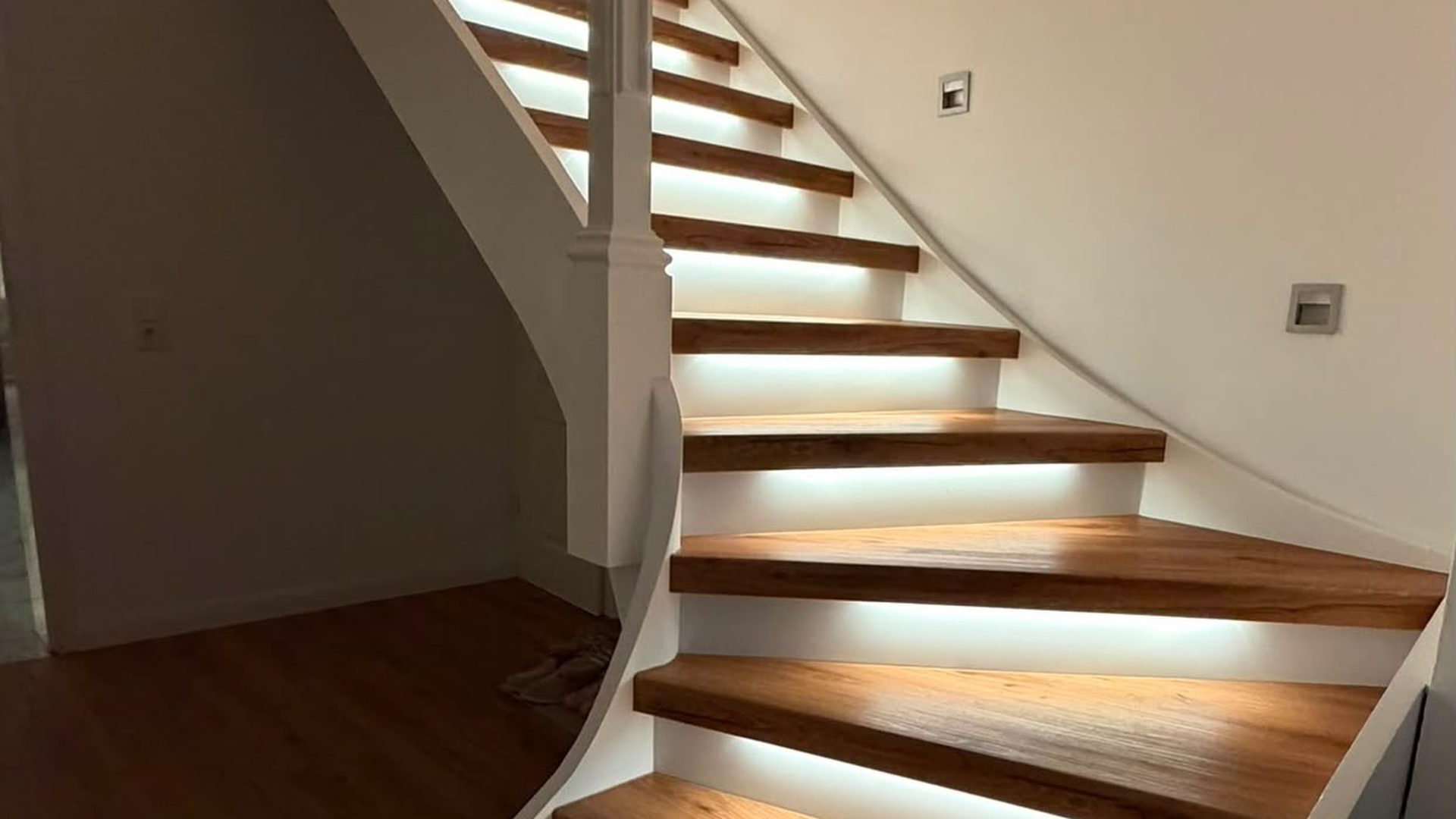 Frisch renovierte, gewendelte Treppe mit warmen Holzstufen, weißen Stellstufen und indirekter LED-Beleuchtung